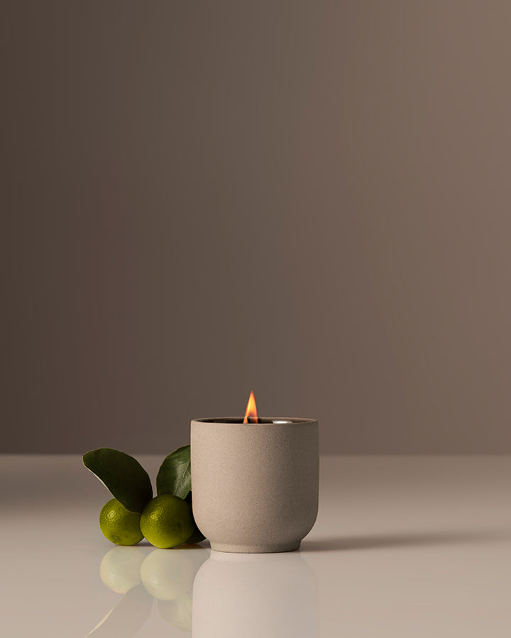 Ceramic candle holder with a lit candle and two limes on a reflective surface.