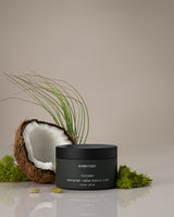 Black jar of Homecourt body butter with a coconut and greenery on a neutral background