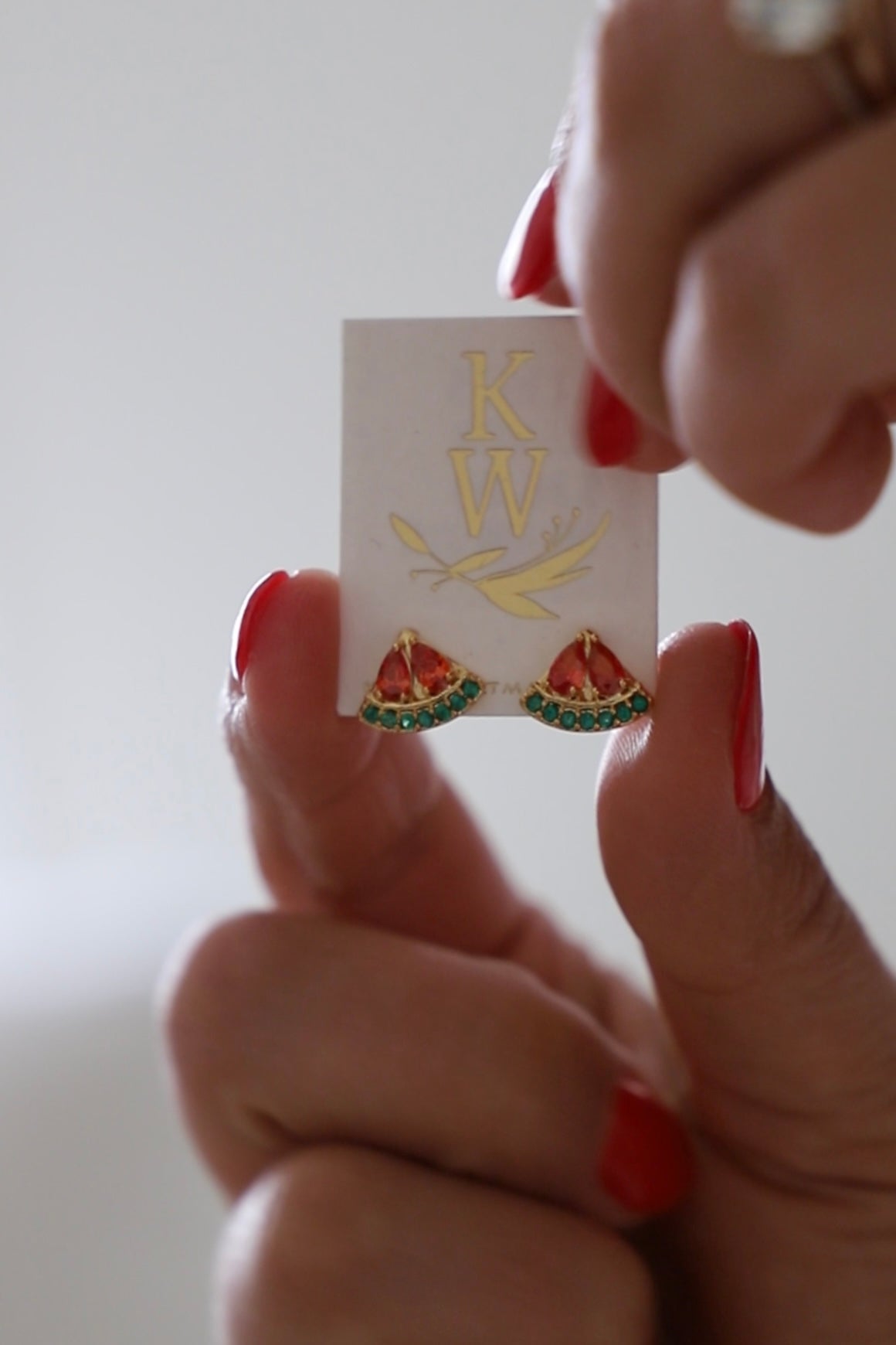 Person holding a card with 'KW' logo and watermelon design earrings against a plain background