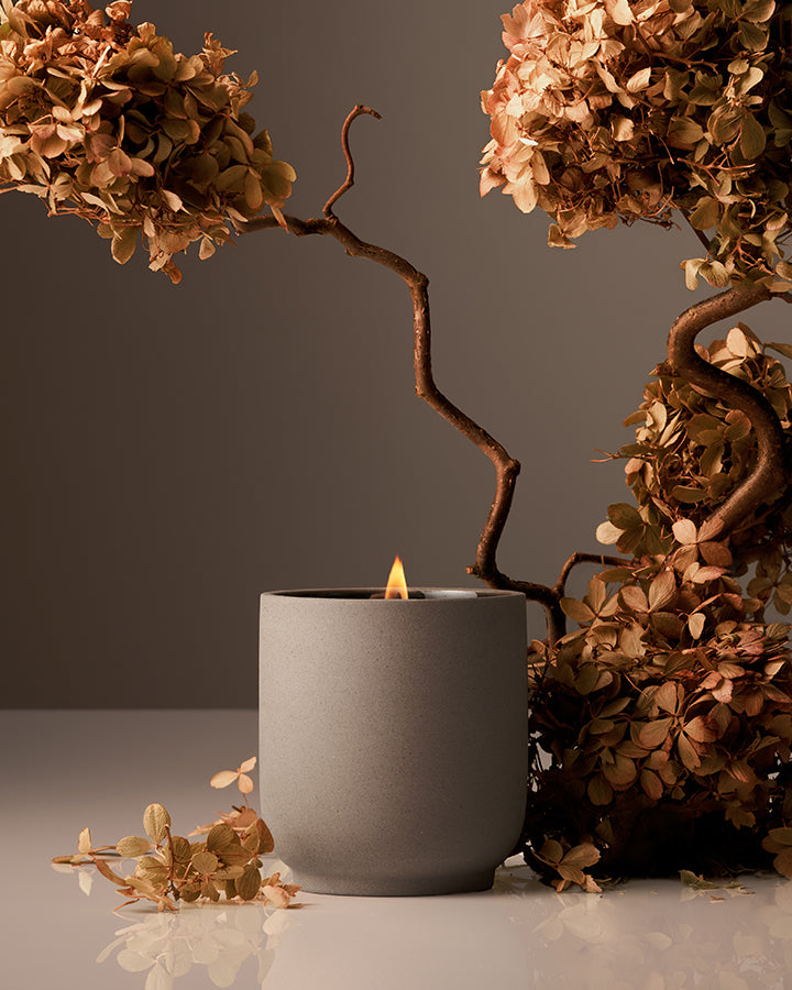 Gray candle in a pot with dried flowers on a reflective surface and dark background