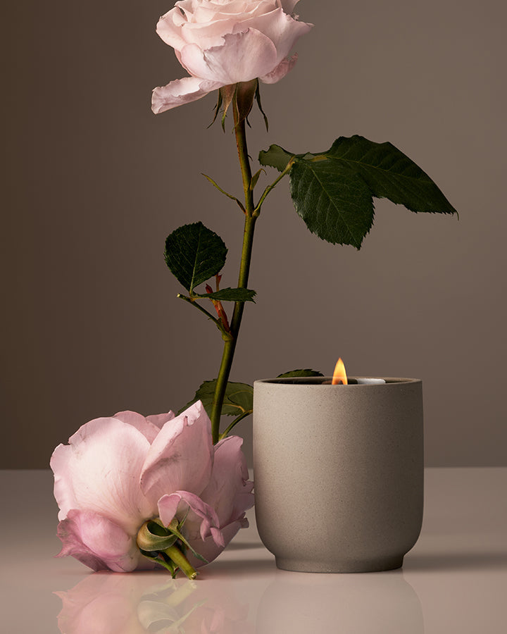 Gray candle holder with a lit candle, pink rose, and green leaves on a reflective surface.