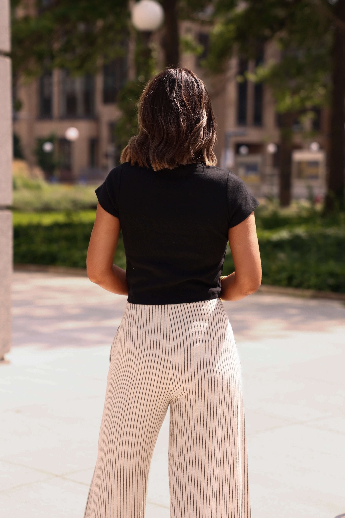 Person wearing a black top and beige pants standing in an outdoor setting with trees and buildings in the background.