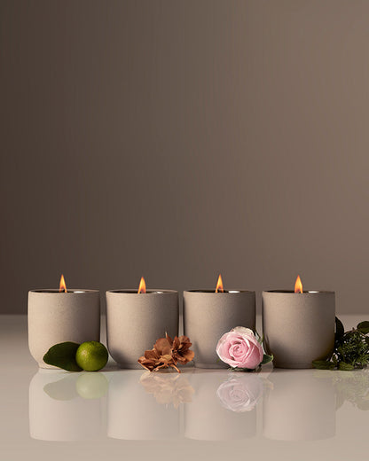 Four lit candles in gray holders on a reflective surface with flowers and leaves.