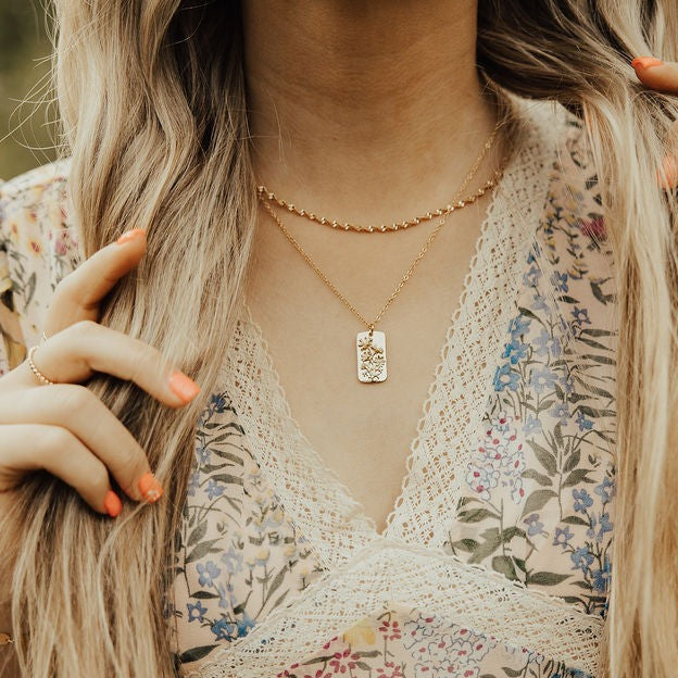 Close-up of a person wearing a gold necklace with a rectangular pendant, set against a floral patterned background.