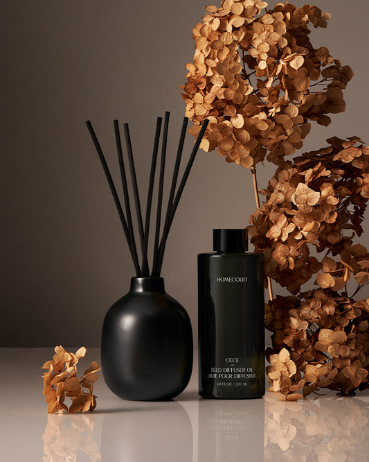 Black diffuser with reeds and a bottle of Home&Garden candle in front of dried flowers on a reflective surface.