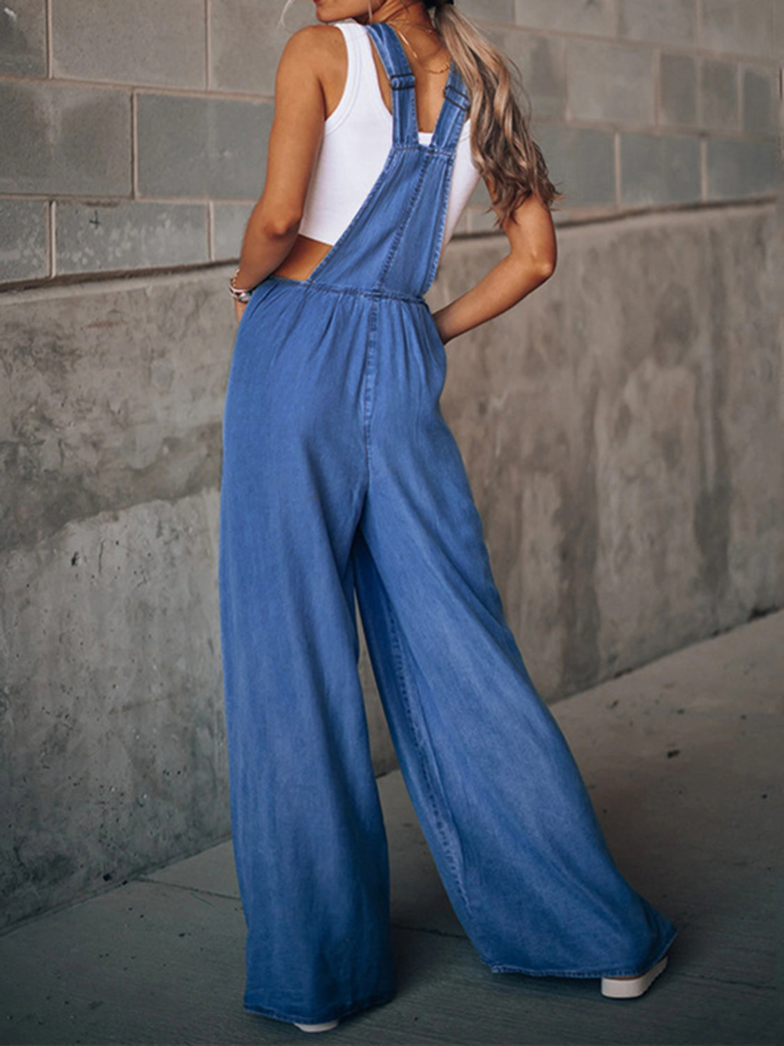 Woman wearing blue wide leg overalls posing near concrete wall
