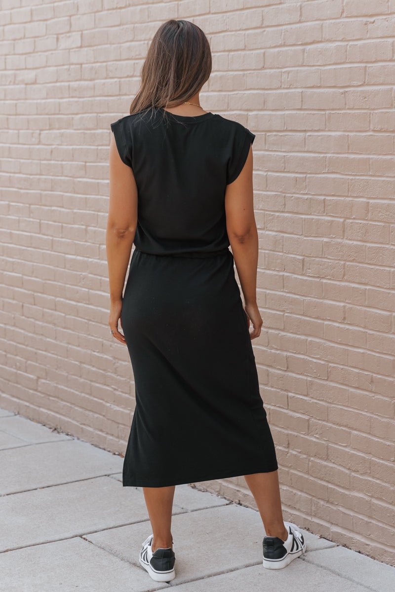 Woman wearing a black dress standing against a beige brick wall.