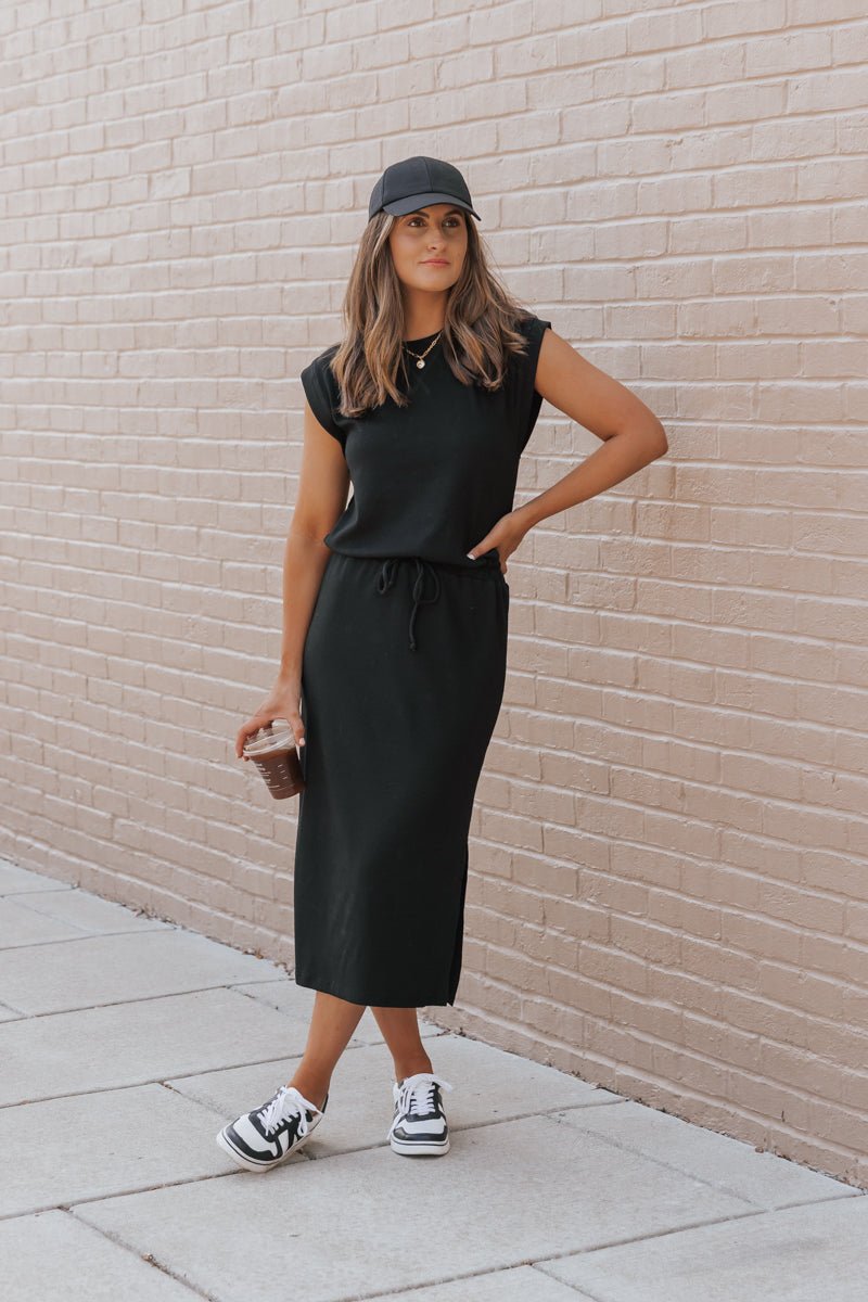 Woman wearing a black dress and cap against a beige brick wall.