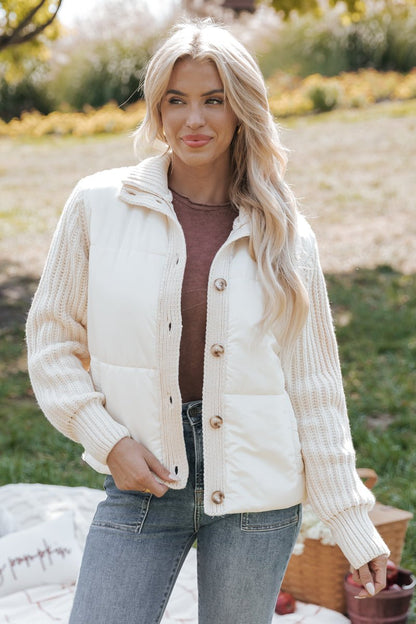 Woman wearing a cream jacket over a brown top and jeans outdoors.