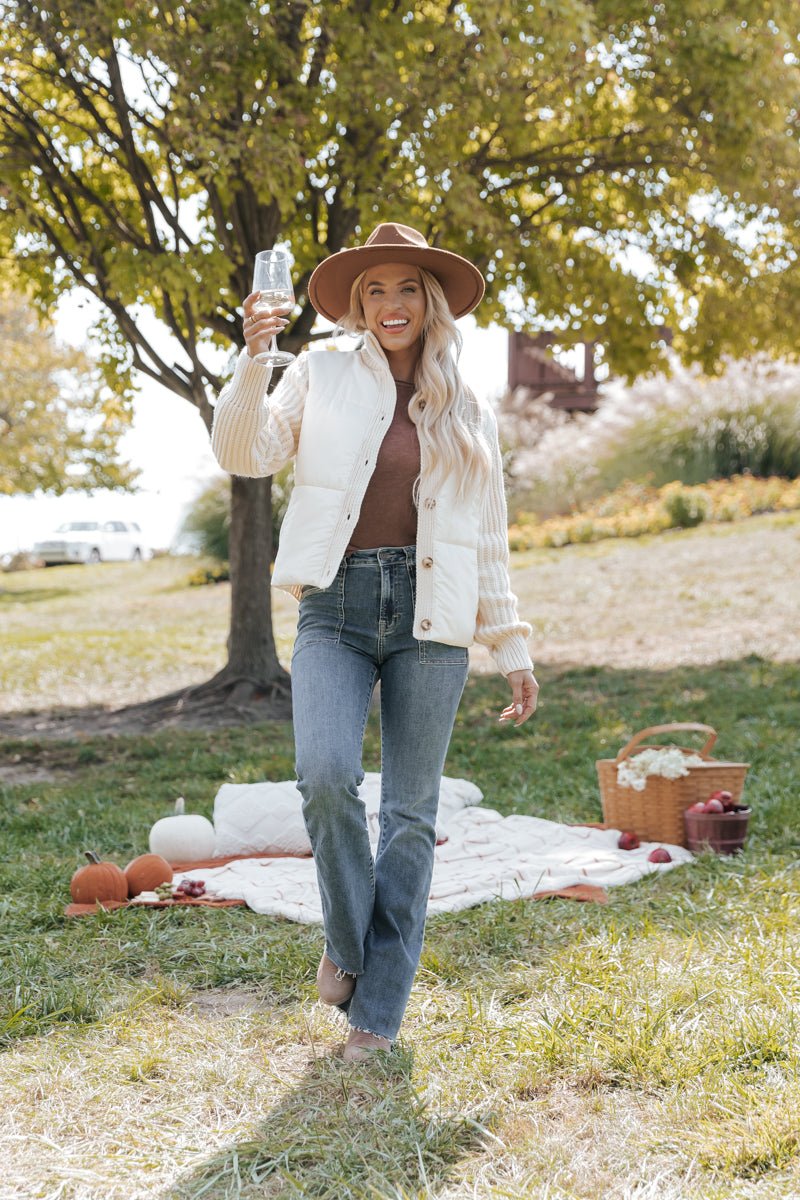 Woman in a park wearing a cream jacket holding a glass of wine with a picnic setup
