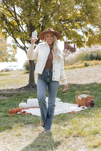 Woman in a park wearing a cream jacket holding a glass of wine with a picnic setup