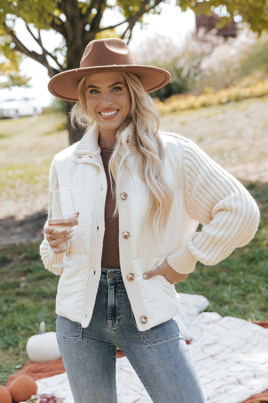 Woman wearing a cream cardigan jacket, brown hat, and jeans, standing outdoors.