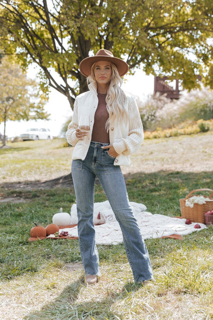 Woman in a park wearing a hat, cream jacket, and jeans with a picnic blanket and basket in the background.