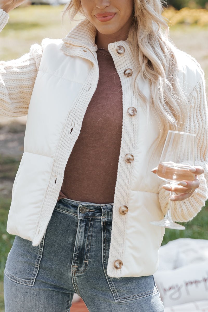 Person wearing a cream-colored knit jacket over a brown top and blue jeans, holding a glass outdoors.