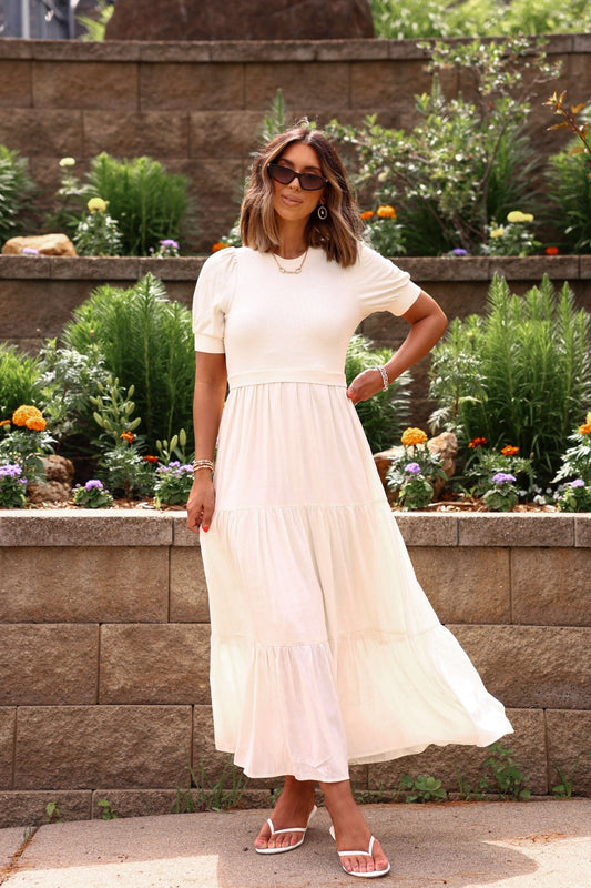 Woman in a cream dress standing in a garden with flowers and plants.