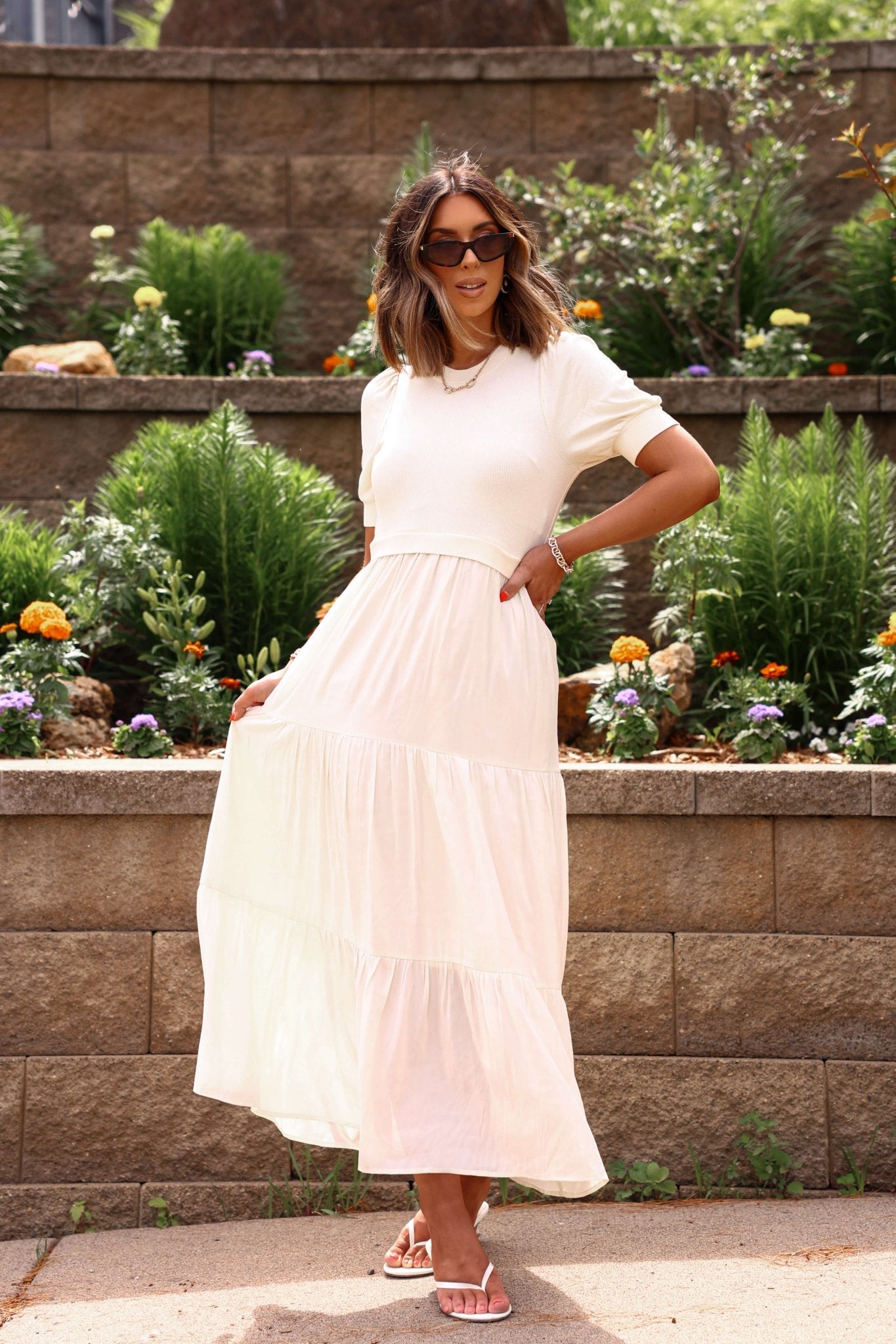 Woman in a white dress standing in front of a garden with plants and flowers.