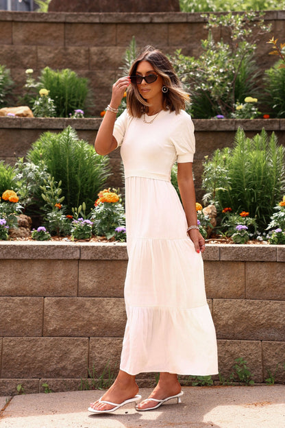 Woman in a white dress standing in front of a garden with flowers and plants.