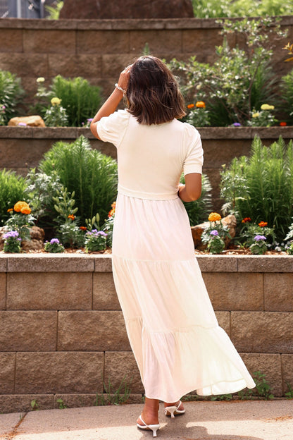 Woman in a white dress standing in front of a garden with plants and flowers.