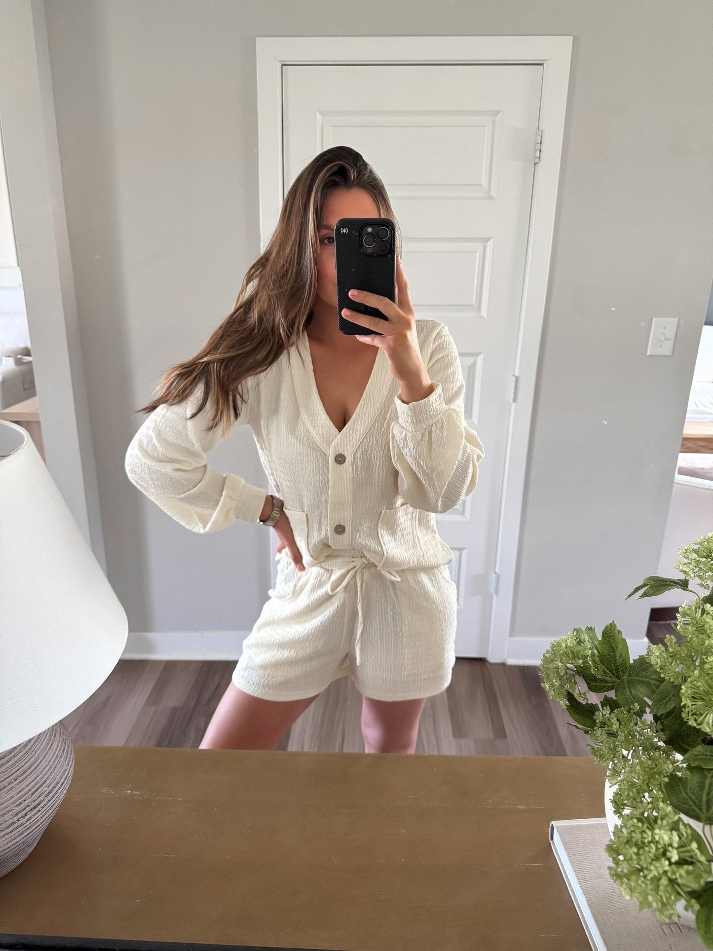 Woman taking a mirror selfie wearing a white cream outfit set in a room with a door and plant.