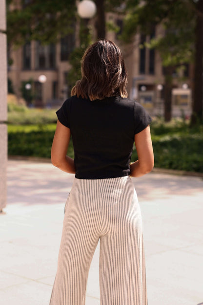Person wearing a black top and beige pants standing in an outdoor setting with trees and buildings in the background.