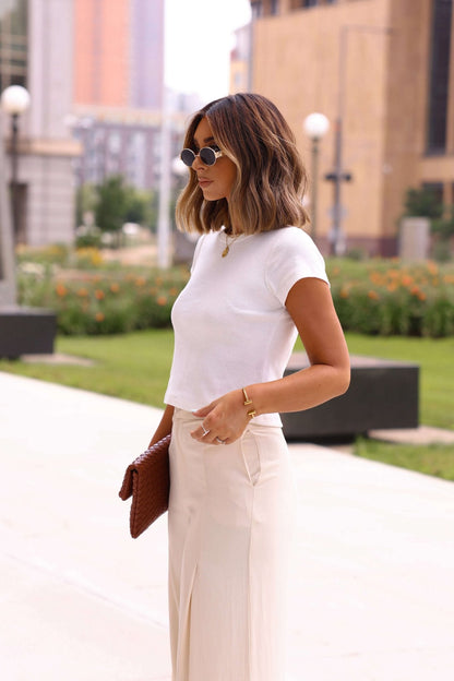 Woman in a white top and beige pants standing outdoors with buildings in the background
