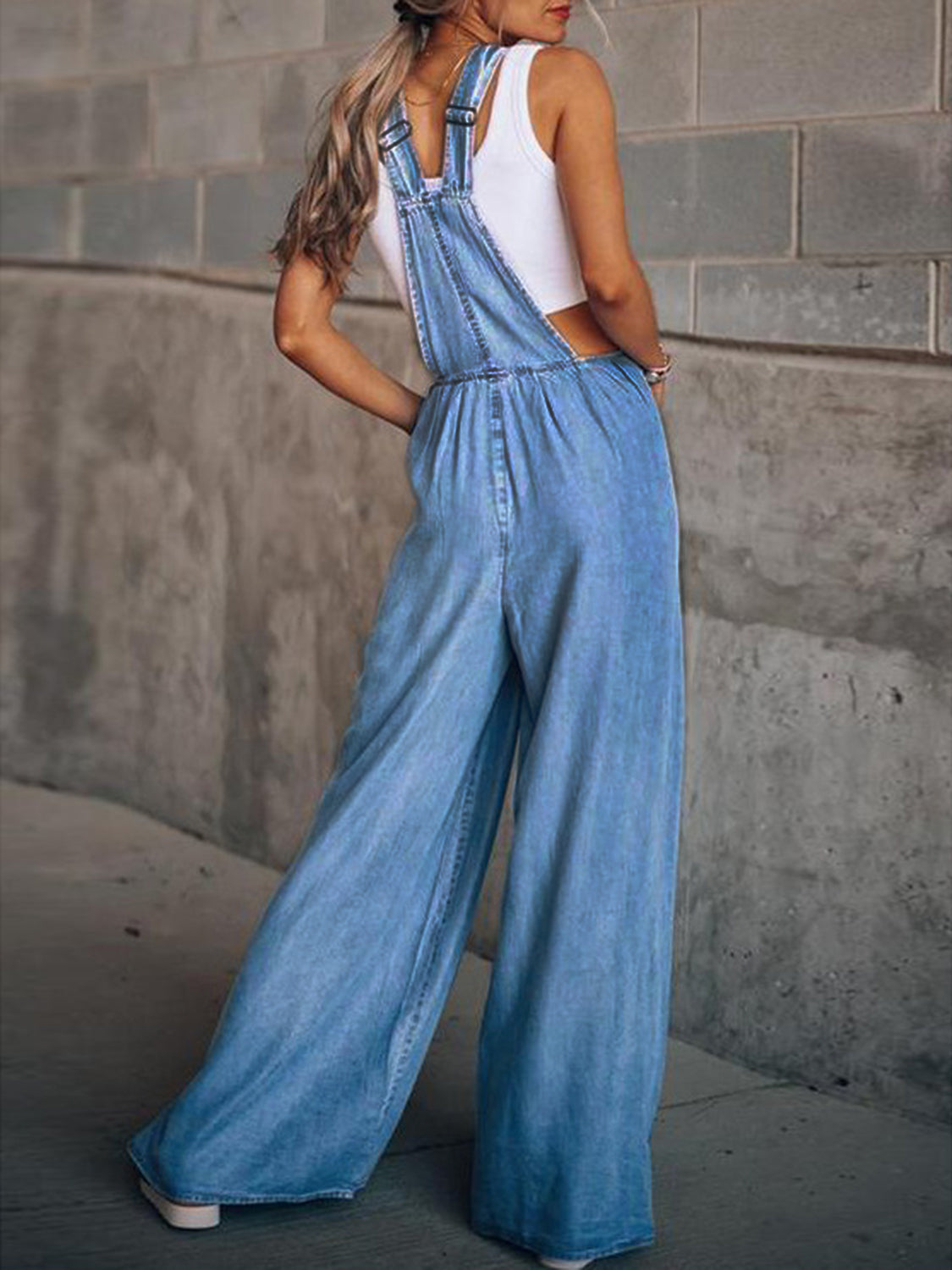 Woman wearing blue wide leg overalls posing against concrete wall